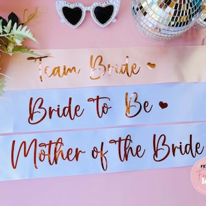 Peut inclure: Trois sashes en satin avec inscription en or. La premi&egrave;re est rose et dit "Team Bride". La deuxi&egrave;me est bleu clair et dit "Bride to Be". La troisi&egrave;me est bleu clair et dit "Mother of the Bride".
