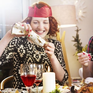 May include: A festive scene with people at a table, opening Christmas crackers. The crackers are white with gold accents. A woman wears a red paper crown and a black and white patterned dress. A lit candle and glasses of red wine are on the table.