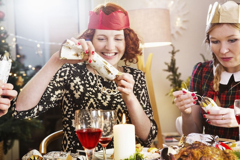 May include: A festive scene with people at a table, opening Christmas crackers. The crackers are white with gold accents. A woman wears a red paper crown and a black and white patterned dress. A lit candle and glasses of red wine are on the table.