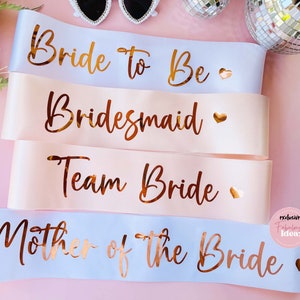 Peut inclure: Quatre ceintures en satin rose et blanc avec des lettres dor&eacute;es. Les ceintures indiquent "Bride to Be", "Bridesmaid", "Team Bride" et "Mother of the Bride".