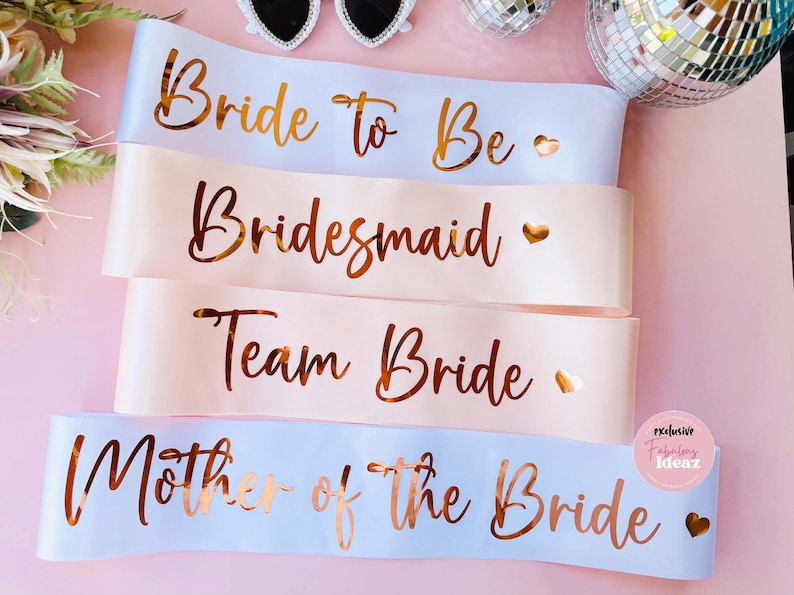 Peut inclure: Quatre ceintures en satin rose et blanc avec des lettres dor&eacute;es. Les ceintures indiquent "Bride to Be", "Bridesmaid", "Team Bride" et "Mother of the Bride".