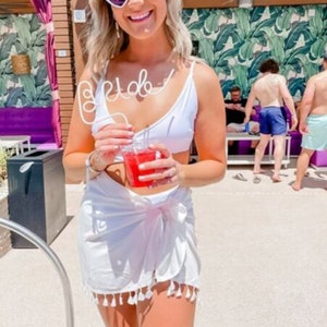 May include: A woman wearing a white swimsuit top and a white sarong with tassels, holding a red drink with a white "Bride" straw. She is wearing heart-shaped sunglasses and smiling.