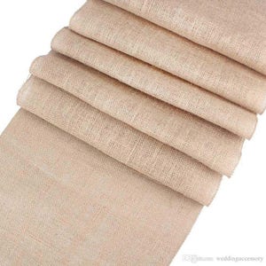 May include: A roll of natural burlap fabric, folded and stacked, with a rough, woven texture. The fabric is a light beige color and is perfect for crafting, decorating, or adding a rustic touch to any project.