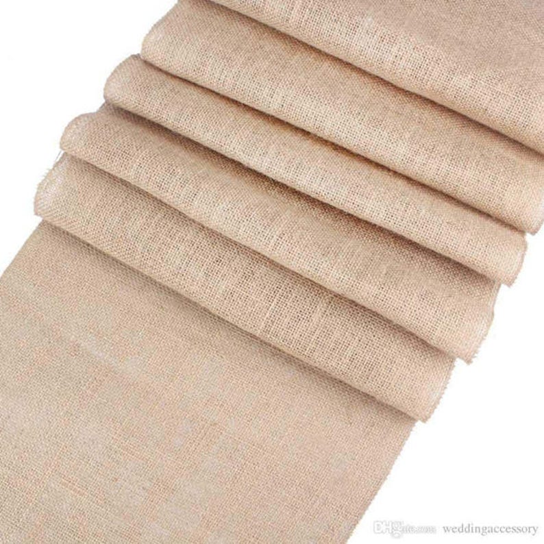 May include: A roll of natural burlap fabric, folded and stacked, with a rough, woven texture. The fabric is a light beige color and is perfect for crafting, decorating, or adding a rustic touch to any project.