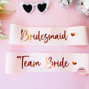 Peut inclure: Deux sashes en satin blanc avec des lettres dor&eacute;es. Le sash du haut dit "Bridemaid" avec un c&oelig;ur dor&eacute;. Le sash du bas dit "Team Bride" avec un c&oelig;ur dor&eacute;.