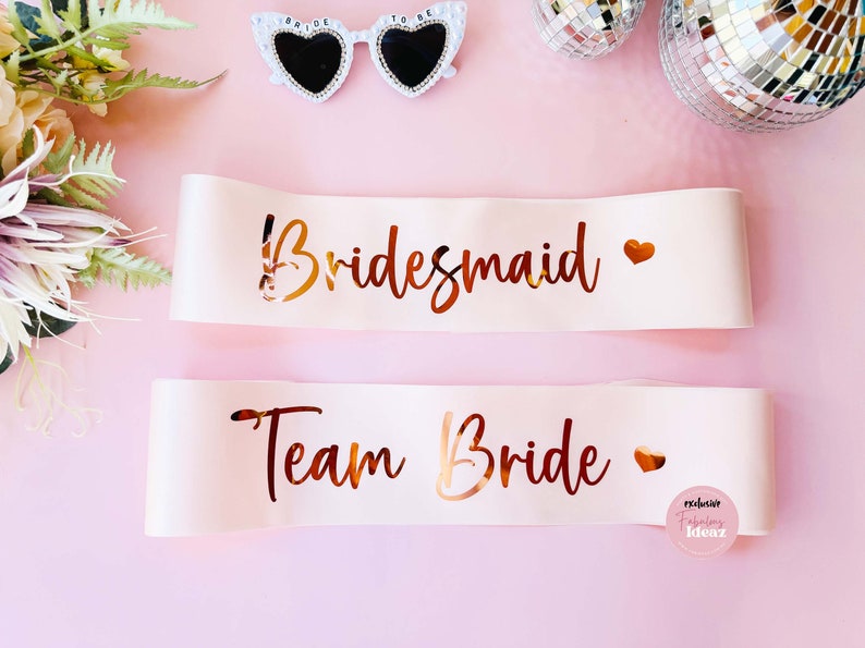 Peut inclure: Deux sashes en satin blanc avec des lettres dor&eacute;es. Le sash du haut dit "Bridemaid" avec un c&oelig;ur dor&eacute;. Le sash du bas dit "Team Bride" avec un c&oelig;ur dor&eacute;.