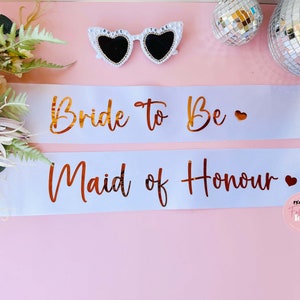 Peut inclure: Deux sashes en satin blanc avec des lettres dor&eacute;es. Le ruban du haut indique "Bride to Be" avec un petit c&oelig;ur. Le ruban du bas indique "Maid of Honour" avec un petit c&oelig;ur.