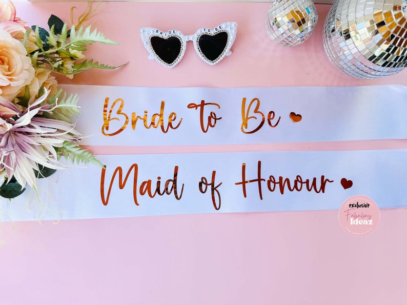 Peut inclure: Deux sashes en satin blanc avec des lettres dor&eacute;es. Le ruban du haut indique "Bride to Be" avec un petit c&oelig;ur. Le ruban du bas indique "Maid of Honour" avec un petit c&oelig;ur.