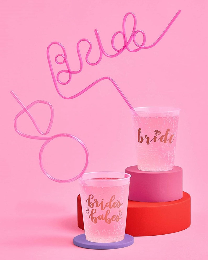 May include: Two clear plastic cups with pink and gold lettering. One cup says "bride" and the other says "bride's babes". A pink bendy straw is shaped to spell "bride" and is in the background.