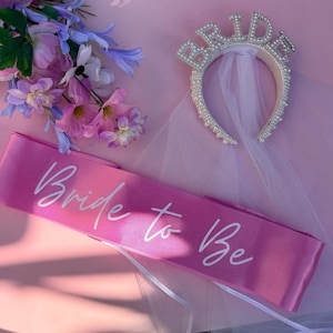 Könnte beinhalten: Ein pinkfarbenes Satinschärpe mit dem Text "Bride to Be" in weißer Schrift, ein mit Perlen und Strasssteinen besetztes Stirnband mit dem Wort "BRIDE" und ein Strauß lila und rosa Blumen. Die Artikel sind auf einer pinkfarbenen Oberfläche angeordnet.