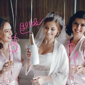 May include: Three friends getting ready for a wedding. The bride is wearing a white robe and holding a bottle of champagne. Her friends are wearing pink robes and holding pink straws that say "bride" and "maid of honor".