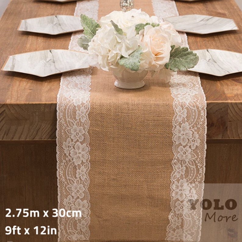 May include: A burlap table runner with white lace trim. The runner is 9 feet long and 12 inches wide. It is shown on a wooden table with white plates and a centerpiece of white and peach flowers.