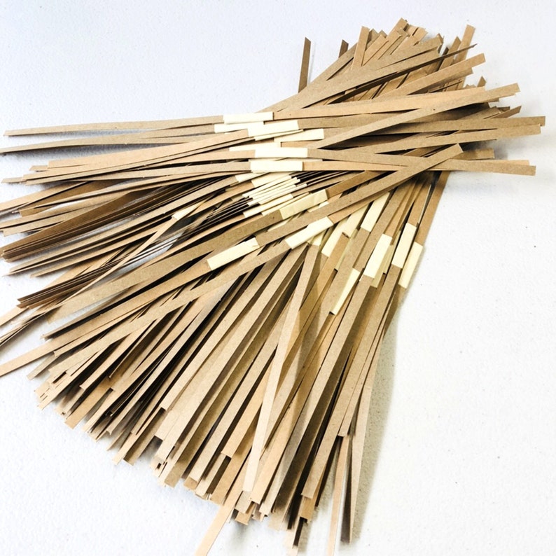 May include: A pile of thin, light brown paper strips, some held together with small, rectangular pieces of tape. The strips are arranged in a slightly fanned-out pattern against a white background. The paper appears to be a natural, unbleached color.