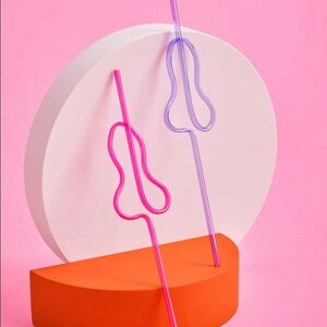 May include: Two colorful, flexible drinking straws with a unique, curved design. The straws are pink and purple and are standing upright on a white and orange platform.