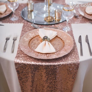 Könnte beinhalten: Eine elegante Tischdekoration mit roségoldenen Akzenten. Ein paillettenbesetzter Tischläufer und Platzteller werden mit einer weißen Tischdecke kombiniert. Eine Serviette wird von einem dekorativen Ring gehalten. Weingläser und Besteck vervollständigen den Look.