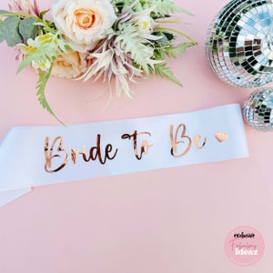 Peut inclure: Sash en satin blanc avec l'inscription "Bride To Be" en lettres dor&eacute;es et un petit c&oelig;ur dor&eacute;.