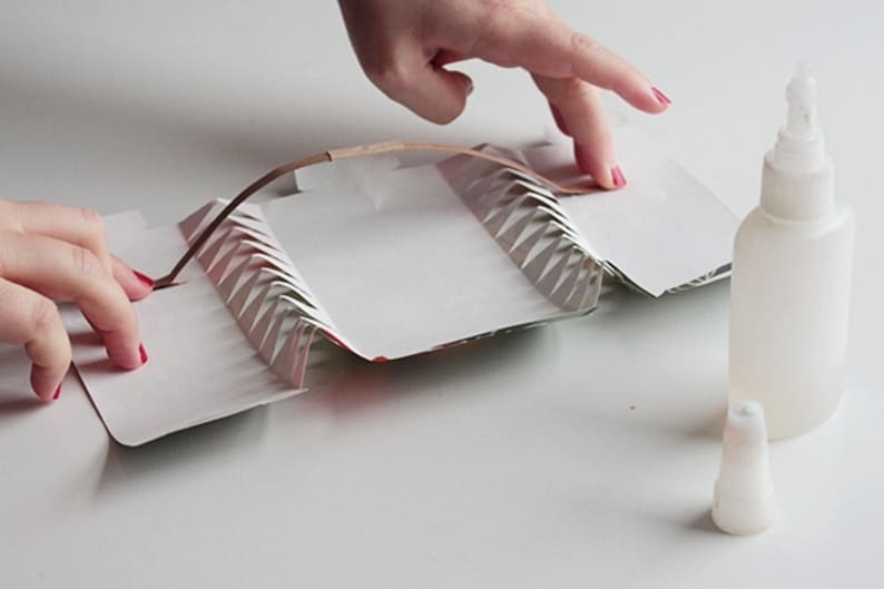 May include: A person's hands are shown assembling a white paper craft with a brown accent. The craft has a zig-zag edge design. A clear glue bottle and cap are on the right side of the image.