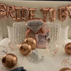 May include: A white bed with rose gold foil balloons spelling "Bride to Be" and a large rose gold ring balloon. There are several rose gold and clear balloons with confetti on the bed, as well as a pink tote bag with the text "Boots & Booze & Besties" and a white hat with the word "Bride" on it.
