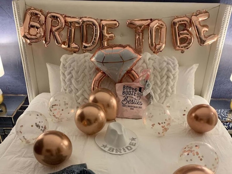 May include: A white bed with rose gold foil balloons spelling "Bride to Be" and a large rose gold ring balloon. There are several rose gold and clear balloons with confetti on the bed, as well as a pink tote bag with the text "Boots & Booze & Besties" and a white hat with the word "Bride" on it.