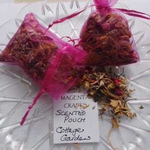 May include: Two pink mesh bags filled with dried flowers and herbs. The bags are on a white plate with a white card that reads "Magenta Crafts Scented Pouch Cottage Garden".