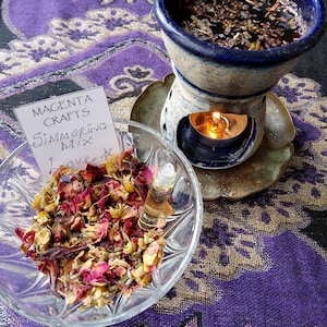 Puede incluir: Un difusor de aceites esenciales de cerámica azul con una vela encendida, un cuenco de vidrio transparente con flores y hierbas secas y un pequeño vial de vidrio. El texto en la tarjeta dice "Magenta Crafts Simmering Mix Love #". Los artículos están sobre una tela morada estampada.