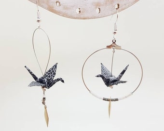 May include: A pair of silver earrings with black and white origami cranes. One earring features a crane hanging from a hoop with a silver bead chain and a gold charm. The other earring features a crane hanging from a silver loop with a gold charm.