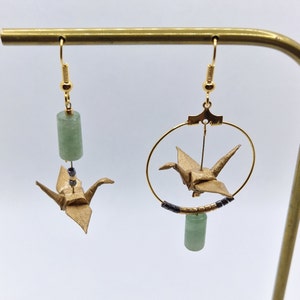 Op de afbeelding: Een paar gouden origami-kraanvogel oorbellen met groene jade-steen accenten. Eén oorbel heeft een hoepel met een kraanvogel die erin hangt, terwijl de andere een kraanvogel heeft die aan een jade-steen hangt.