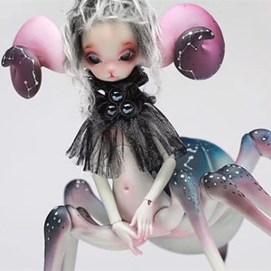 May include: A fantasy art doll with a spider-like body and a human-like torso. The doll has a pale complexion, large eyes, and gray hair. It wears a black ruffled collar and has large, pink ear-like appendages with dark purple accents.