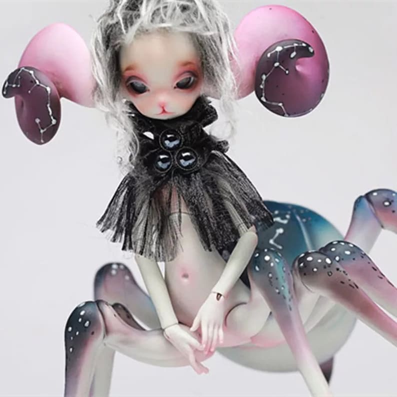 May include: A fantasy art doll with a spider-like body and a human-like torso. The doll has a pale complexion, large eyes, and gray hair. It wears a black ruffled collar and has large, pink ear-like appendages with dark purple accents.