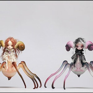 May include: Two fantasy dolls with spider-like bodies and doll faces. One doll has red hair and large, curved horns. The other doll has gray hair and pink ear-like structures. Both dolls have multiple legs and are set against a white background.