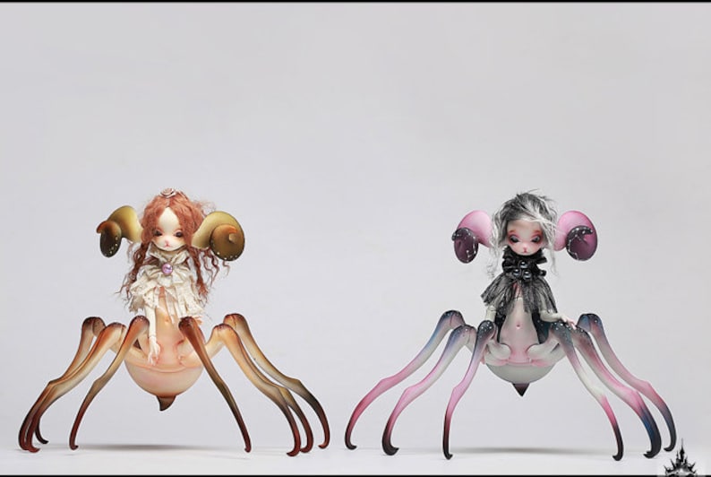 May include: Two fantasy dolls with spider-like bodies and doll faces. One doll has red hair and large, curved horns. The other doll has gray hair and pink ear-like structures. Both dolls have multiple legs and are set against a white background.