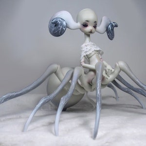 May include: A unique, pale-skinned doll with a spider-like body and a delicate, human-like torso. The doll has large, curved ears with dark blue accents and is dressed in a white lace top. The legs are long and gray, with subtle blue veining.