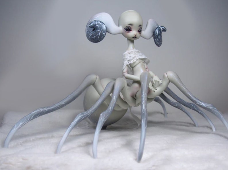 May include: A unique, pale-skinned doll with a spider-like body and a delicate, human-like torso. The doll has large, curved ears with dark blue accents and is dressed in a white lace top. The legs are long and gray, with subtle blue veining.