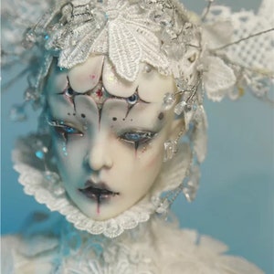 May include: A detailed close-up of a doll's face with elaborate white lace and crystal embellishments. The doll has pale skin, dark eye makeup, and a dark lip color. The doll's head is adorned with a white lace headpiece.