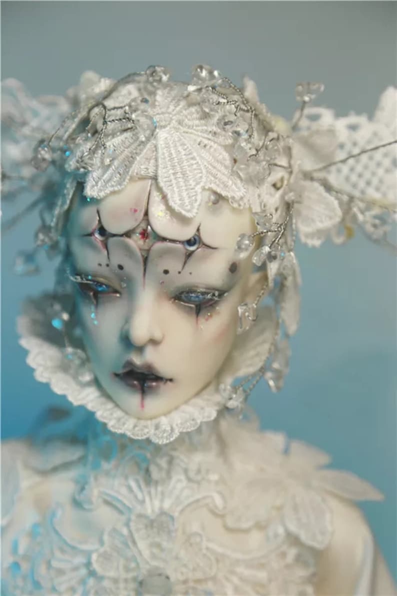 May include: A detailed close-up of a doll's face with elaborate white lace and crystal embellishments. The doll has pale skin, dark eye makeup, and a dark lip color. The doll's head is adorned with a white lace headpiece.