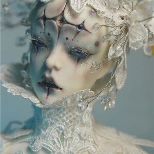 May include: A detailed close-up of a doll's face with pale skin, blue eyes, and elaborate white lace and crystal embellishments. The doll has dark eye makeup and a delicate, ornate headpiece. The overall aesthetic is ethereal and artistic.