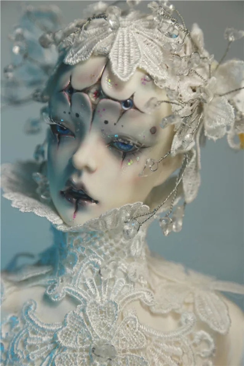 May include: A detailed close-up of a doll's face with pale skin, blue eyes, and elaborate white lace and crystal embellishments. The doll has dark eye makeup and a delicate, ornate headpiece. The overall aesthetic is ethereal and artistic.