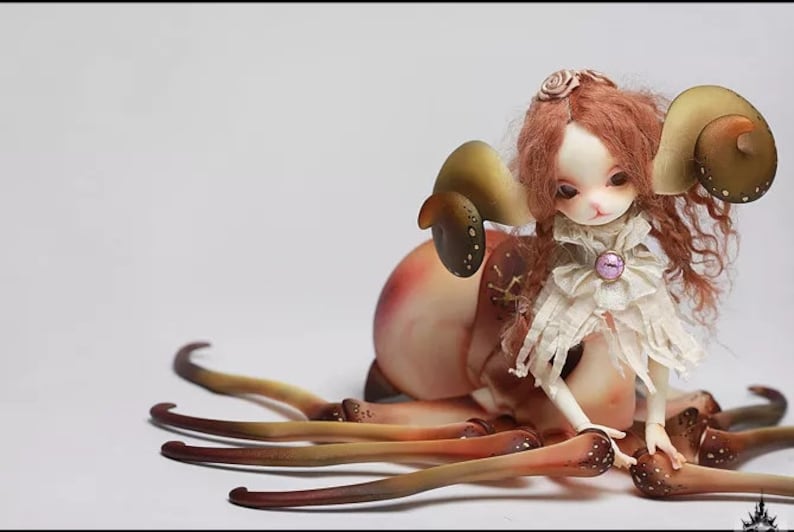 May include: A detailed doll with a unique design. The doll features a pale complexion, red hair, and large, curved, horn-like structures. It wears a white, ruffled outfit and is surrounded by multiple brown tentacles. A pink gem adorns the doll's chest.
