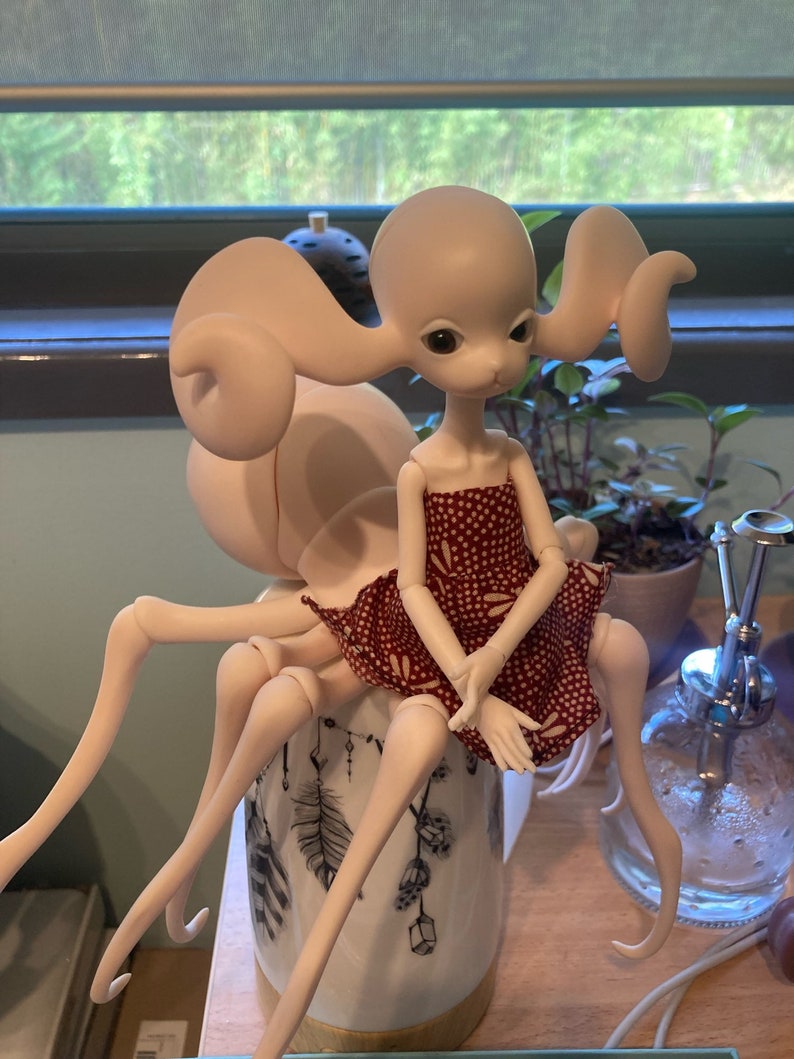 May include: A unique doll with a pale complexion and large eyes, wearing a red dress with white polka dots. The doll has an unusual body shape, with multiple slender limbs and a large, rounded form behind its head. It sits atop a decorative ceramic pot.