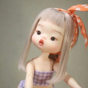 May include: A doll with a pale complexion, gray hair, and large dark eyes. The doll wears a purple gingham top and a matching headband with white polka dots on an orange background. The doll's lips are slightly parted, and it has a neutral expression.