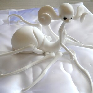 May include: A white, articulated doll with an insect-like body and a humanoid head. The doll has long, slender limbs and a curved torso. Its head features large, dark eyes and stylized, curled antennae. The doll is posed on a white surface.