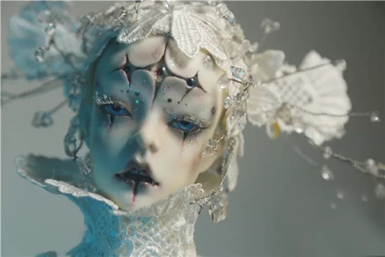 May include: A detailed close-up of a doll's face with pale skin, blue eyes, and dark accents around the eyes and mouth. The doll wears a white, ornate headpiece with floral and crystal embellishments, creating a fantasy or gothic aesthetic.
