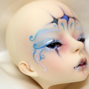 May include: A pale-skinned doll head with detailed facial features. The doll has blue and white painted designs around the eyes and forehead, with long white eyelashes. The lips are a pale pink, and the eyes are a deep blue.