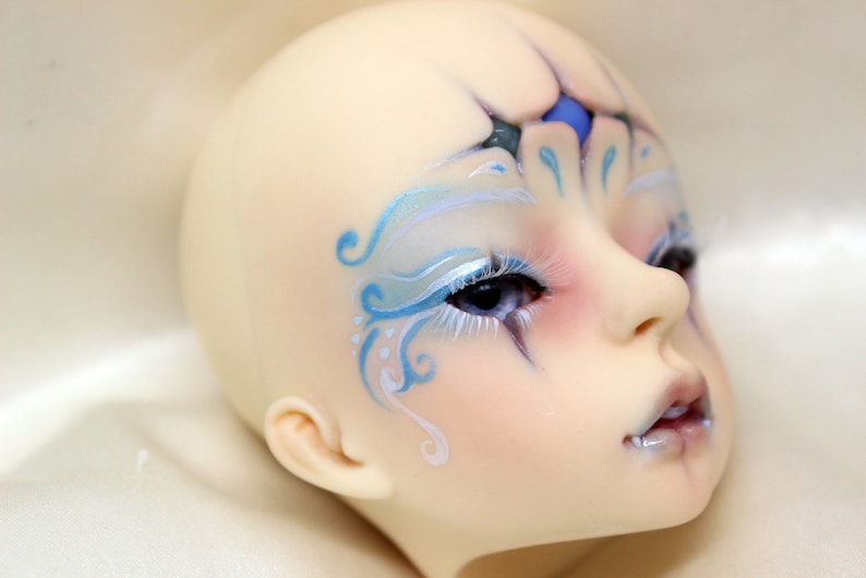 May include: A pale-skinned doll head with detailed facial features. The doll has blue and white painted designs around the eyes and forehead, with long white eyelashes. The lips are a pale pink, and the eyes are a deep blue.