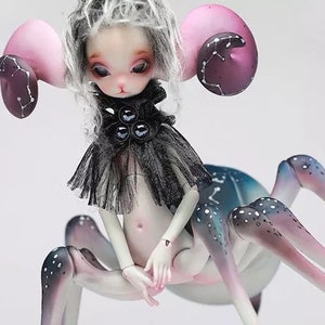 May include: A fantasy art doll with a spider-like body and a human-like torso. The doll has pale skin, large pink ears with dark purple accents, and a black and gray outfit. The legs are a gradient of pink, blue, and white with white spots.