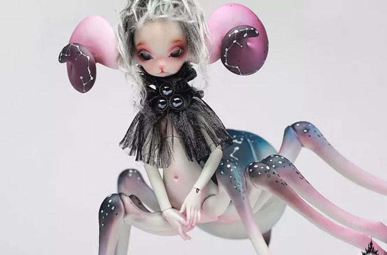 May include: A fantasy art doll with a spider-like body and a human-like torso. The doll has pale skin, large pink ears with dark purple accents, and a black and gray outfit. The legs are a gradient of pink, blue, and white with white spots.