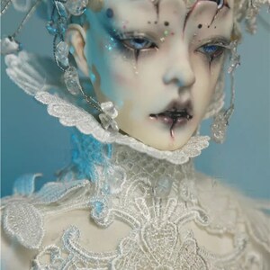 May include: A detailed close-up of a doll with pale skin, blue eyes, and dark makeup. The doll wears a white lace collar and headpiece adorned with crystals. The overall aesthetic is gothic and ethereal.