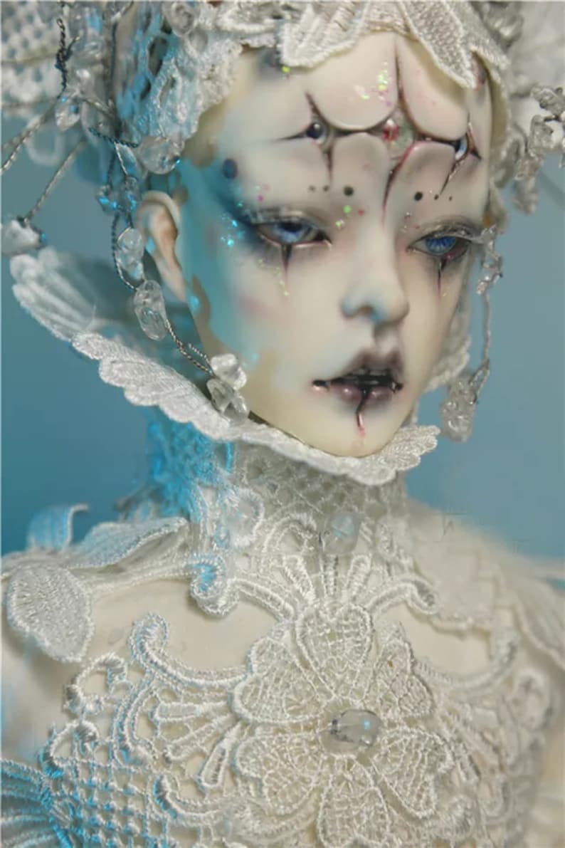 May include: A detailed close-up of a doll with pale skin, blue eyes, and dark makeup. The doll wears a white lace collar and headpiece adorned with crystals. The overall aesthetic is gothic and ethereal.