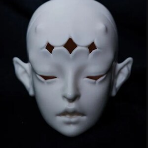 May include: A white ceramic doll face with closed eyes and small, pointed ears. The forehead features a decorative pattern of diamond-shaped cutouts. The face has a smooth, matte finish, and the overall design is stylized and artistic.