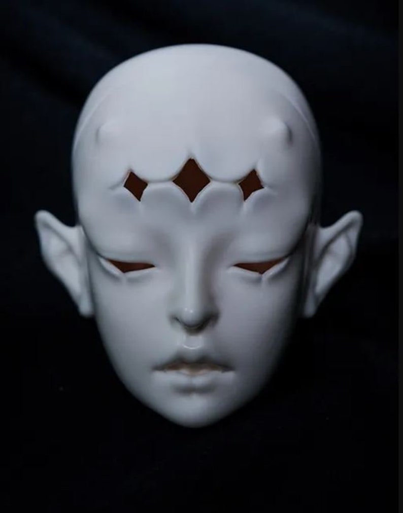 May include: A white ceramic doll face with closed eyes and small, pointed ears. The forehead features a decorative pattern of diamond-shaped cutouts. The face has a smooth, matte finish, and the overall design is stylized and artistic.
